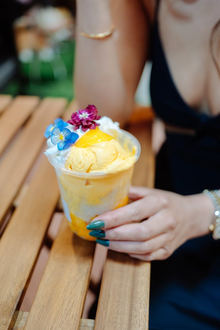 Colorful mango dessert with vibrant flowers on a wooden table, held by a woman.