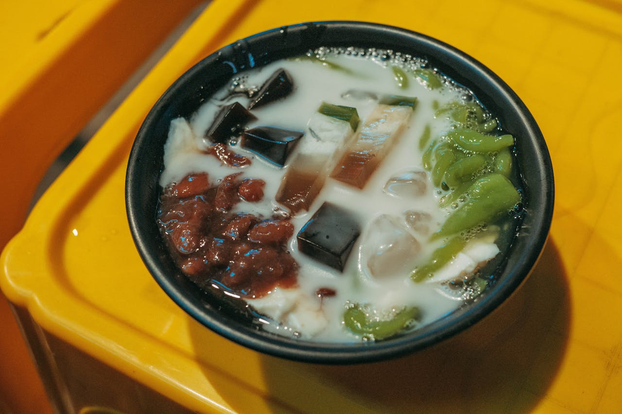 hero-img-01 A delicious bowl of Asian dessert featuring colorful jelly cubes, red beans, and creamy milk on a vibrant yellow background.