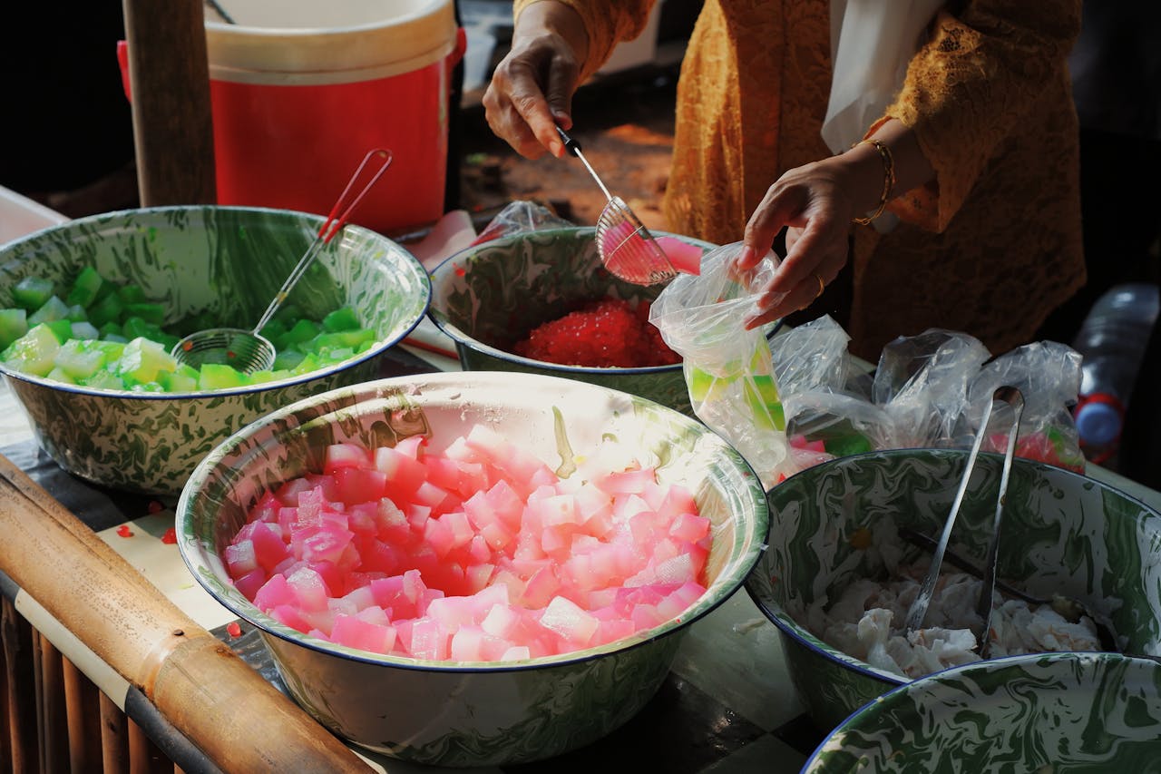 hero-img-02 Woman preparing traditional Indonesian dessert with colorful ingredients at an outdoor market.